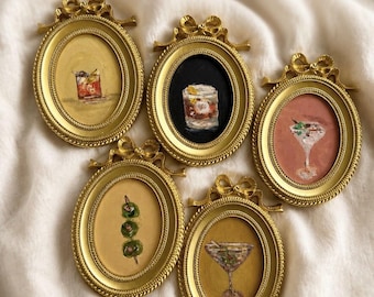 Op de afbeelding: Vijf ovale gouden lijsten met sierlijke strikdetails aan de bovenkant, elk met een klein schilderij van een cocktail. De schilderijen tonen verschillende drankjes, waaronder een glas met olijven, een martini en andere mixdrankjes op gekleurde achtergronden.