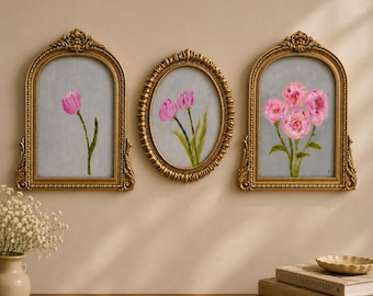 Op de afbeelding: Drie wandkunstwerken in sierlijke gouden lijsten. Twee lijsten zijn boogvormig met decoratieve bovenkanten en één is ovaal. Elk bevat een schilderij van roze bloemen op een lichtgrijze achtergrond. De lijsten hebben gedetailleerde reliëfs.