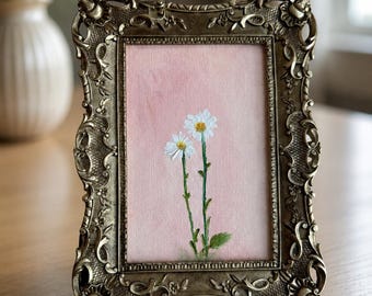 Op de afbeelding: Een rechthoekige fotolijst met een sierlijke, antiek bronskleurige metalen rand met ingewikkelde krullen en bloemmotieven. Binnenin toont een schilderij op een zachtroze achtergrond twee witte madeliefjes met gele harten.
