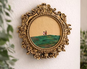 Op de afbeelding: Omlijst kunstwerk in sierlijk goud. Het ronde schilderij toont twee figuren op een groene heuvel tegen een gouden achtergrond. De lijst heeft een uitgebreid ontwerp en is op een witte muur gemonteerd.