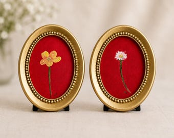 Op de afbeelding: Twee ovale gouden lijsten met kralenranden, elk met een rode stoffen achtergrond. De linkerlijst toont een schilderij van een gele bloem, terwijl de rechterlijst een schilderij van een witte bloem bevat. Beide lijsten staan op zwarte standaards.