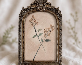 Op de afbeelding: Een boogvormige fotolijst met een sierlijke, antiek goudkleurige metalen rand met ingewikkeld krulwerk en bloemmotieven. Binnenin toont een lichtbeige, getextureerde achtergrond een delicate illustratie van twee gekruiste bloemstengels.