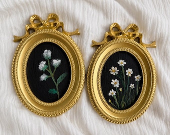 Op de afbeelding: Twee ovale wandlijsten met een metallic gouden afwerking en decoratieve strikaccenten aan de bovenkant. Elke lijst bevat een zwarte achtergrond met handgeschilderde witte bloemen en groene stelen. De lijsten hebben een textuur met kraalrand.