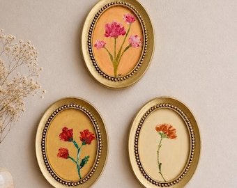 Op de afbeelding: Drie ovale wanddecoraties in gouden kralenlijsten. De bovenste lijst toont roze bloemen op een oranje achtergrond. De linkeronderlijst toont rode bloemen op een gouden achtergrond. De rechteronderlijst toont een enkele oranje bloem op een crèmekleurige achtergrond.