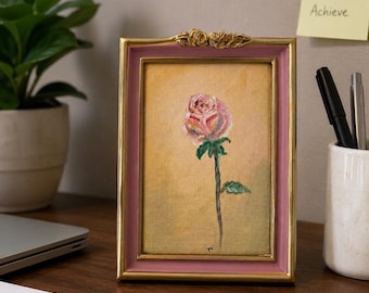 Op de afbeelding: Een rechthoekig ingelijst olieverfschilderij met een enkele roze roos met groene bladeren op een zachte gele achtergrond. De lijst is goudkleurig met een roze binnenrand en een decoratief bloemaccent aan de bovenzijde. Gesigneerd met initialen.
