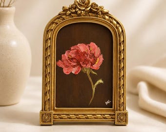 Op de afbeelding: Een boogvormige, goudkleurige fotolijst met sierlijke roosgravures aan de bovenkant en bloemmotieven aan de voet. De lijst bevat een klein olieverfschilderij van een enkele roze pioenroos op een donkerbruine achtergrond, met de initialen MV.