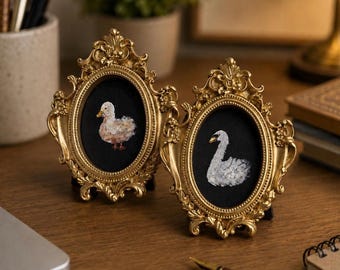 Op de afbeelding: Twee ovale fotolijsten met sierlijke goudkleurige randen. Elke lijst bevat een klein schilderij op een zwarte achtergrond, de ene met een lichtgekleurd eendenkuiken en de andere met een witte zwaan. De lijsten hebben ingewikkelde krulmotieven.