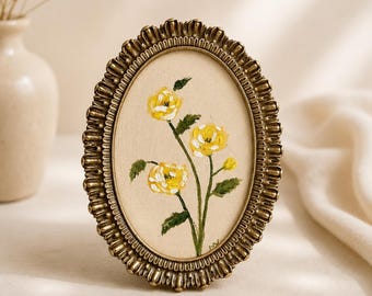 Op de afbeelding: Een ovaalvormige fotolijst met een metallic bronzen afwerking en een gegroefde rand, met een met stof beklede inzetstuk voorzien van een handgeschilderde illustratie van drie gele bloemen met groene bladeren en stengels op een lichtbeige achtergrond.