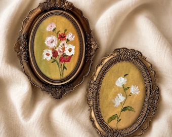 Op de afbeelding: Twee ovale wandlijsten met sierlijke, donkerbronskleurige metalen randen. De linkerlijst bevat een schilderij van een kleurrijk bloemenboeket met witte, roze en rode bloemen. De rechterlijst toont een schilderij van drie witte bloemen.