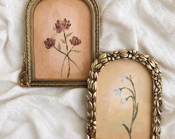 Op de afbeelding: Twee boogvormige goudkleurige lijsten met botanische illustraties. De linkerlijst bevat drie donkerroze bloemen op een perzikkleurige achtergrond. De rechterlijst heeft een bloemmotief en toont witte klokvormige bloemen op een beige achtergrond.