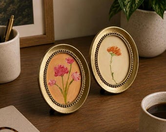 Op de afbeelding: Twee ovale gouden lijsten met kralenranden, elk met een klein bloemenschilderij. De linkerlijst toont een tros roze bloemen op een perzikkleurige achtergrond, terwijl de rechterlijst een enkele oranje bloem op een crèmekleurige achtergrond bevat.