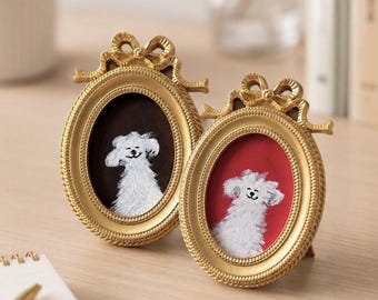 Op de afbeelding: Twee ovale, goudkleurige fotolijsten met decoratieve strikaccenten aan de bovenkant. Elke lijst bevat een klein, wit, gestructureerd schilderij van een hond. De ene lijst heeft een donkerbruine achtergrond en de andere een effen rode achtergrond.