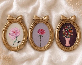 Op de afbeelding: Drie ovale goudkleurige lijsten met decoratieve strikken aan de bovenkant. Elke lijst bevat een klein schilderij: een roze roos op een crèmekleurige achtergrond, een rode roos op een lavendelkleurige achtergrond en een boeket bloemen in een vaas.
