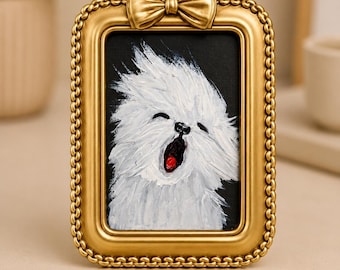 Op de afbeelding: Een rechthoekige goudkleurige fotolijst met een kralenrand en een decoratieve strik aan de bovenkant. De lijst bevat een olieverfschilderij van een pluizige witte hond die met open bek gaapt, tegen een effen zwarte achtergrond.