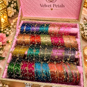 May include: A gold-toned storage box with a pink interior lining and the text Velvet Petals, containing rows of multi-colored glass bangles. Each bangle features small gold-toned metallic accents and beads arranged in a repeating pattern.