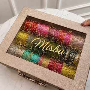 May include: A rose gold glitter box with a clear lid, filled with colorful bangles. The bangles are in shades of red, pink, green, and gold. The name "Misba" is written in gold script across the top of the bangles.