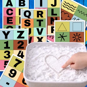 Könnte beinhalten: Bunte Lernkarten mit Buchstaben, Zahlen und Formen. Ein Kinderfinger zeichnet ein Herz in eine weiße, strukturierte Substanz. Geeignet für frühes Lernen und pädagogische Aktivitäten.