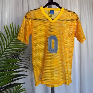 以下が含まれることがあります： Vネックと半袖のイエローメッシュジャージー。ジャージーの前面にはデニムカラーの数字「0」のパッチが付いています。ジャージーは黒いハンガーに吊るされています。