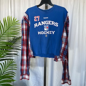 May include: Blue cropped sweatshirt with "Rangers Hockey" text and the number 27. The sleeves are made of red, white, and blue plaid fabric. The shirt is hanging on a black hanger.