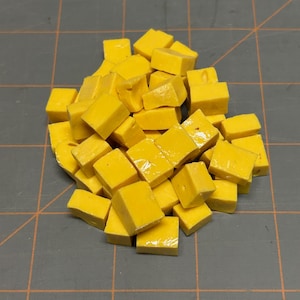 May include: A pile of small, yellow, square-shaped objects. The objects appear to be made of a solid material with a slightly glossy finish. They are scattered on a gray surface with a grid pattern.