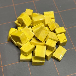 May include: A pile of small, yellow, square-shaped mosaic tiles. The tiles are a solid, bright yellow color and appear to be made of glass or a similar material. They are scattered on a gray surface.