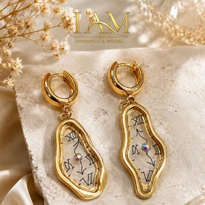 May include: A pair of gold-toned hoop earrings featuring irregular, melted-style frames. Each earring contains a clear center panel printed with black Roman numerals and clock hands, accented by a single iridescent crystal at the center point.