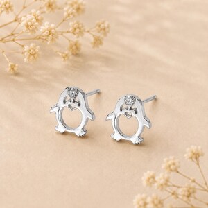 Puede incluir: Un par de pendientes de botón en forma de pingüino de tono plateado. Cada pendiente presenta un diseño de corazón recortado en el centro del cuerpo del pingüino y una pequeña piedra preciosa redonda y transparente en la parte superior.