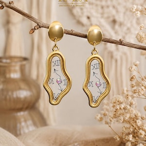 May include: A pair of gold-toned drop earrings featuring an irregular, melted-style frame. The centre of each earring is clear with black Roman numerals and clock hands printed on the surface, accented by a small, iridescent circular stone.