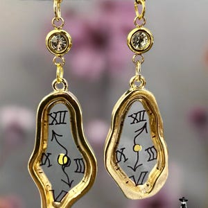 Op de afbeelding: Een paar goudkleurige oorbellen met onregelmatige, gesmolten klokvormen, zwarte Romeinse cijfers en gele stippen in het midden. Elke oorbel hangt aan een kleine gouden schakel verbonden met een ronde knop met een heldere kristal.