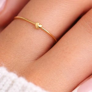 May include: A thin gold ring featuring a small, polished heart-shaped charm centred on the band. The ring has a smooth, metallic finish and a delicate, slender profile that encircles the finger, showcasing a simple and refined design.
