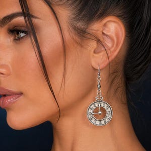 May include: A silver-toned drop earring featuring a circular clock face design with black numerals from one to twelve. The earring hangs from a decorative metal link and a standard hook wire, showcasing a detailed miniature timepiece aesthetic.