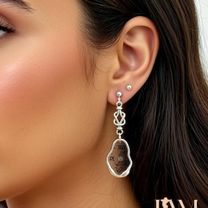 May include: Silver-toned dangle earrings. The earrings feature a series of geometric shapes and a teardrop-shaped charm with Roman numerals. The earrings are worn on a person's ear.