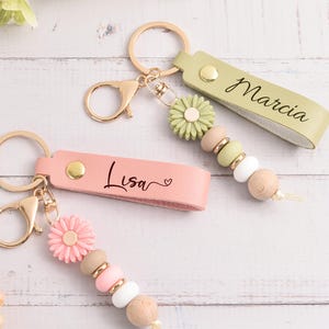 May include: Two personalized keychains with gold-colored hardware. One is light pink with "Lisa" printed on it, and the other is light green with "Marcia" printed on it. Both have daisy-shaped charms and beaded accents in various colors.