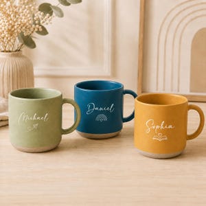 Pode incluir: Três canecas de cerâmica em verde sálvia, azul profundo e amarelo mostarda, cada uma com uma alça e uma base cinza claro contrastante. As canecas são personalizadas com os nomes Michael, Daniel e Sophia em escrita branca, acompanhadas por pequenos ícones de linha de um avião de papel, um arco-íris e um livro aberto.