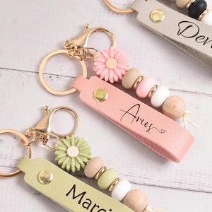 Op de afbeelding: Sleutelhangers met goudkleurige sluitingen en leren bandjes in roze, groen en grijs. Elke sleutelhanger heeft een naam of sterrenbeeld, decoratieve kralen en een bloemenbedel. De middelste sleutelhanger heeft de naam "Aries".