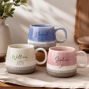 Puede incluir: Tres tazas de cerámica con base moteada y una sección superior lisa y coloreada. La taza azul lleva el nombre Daniel, la taza verde claro lleva el nombre William con un icono de arcoíris y la taza rosa lleva el nombre Sophia con un icono de hoja.