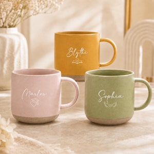 May include: Three ceramic mugs in pastel pink, mustard yellow, and sage green. Each mug features a white handle and a base with a speckled texture. The pink mug says Marlow with a graduation cap, yellow says Blythe with a book, and green says Sophia with wings.