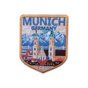 May include: Embroidered patch featuring a shield design with the text "MUNICH GERMANY." The patch depicts a cityscape with two towers, red rooftops, and a mountain range in the background. The patch has a gold border.