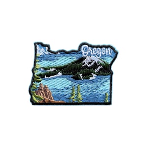 Puede incluir: Parche bordado con la forma de Oregón, que representa un paisaje con agua azul, árboles verdes y una montaña. La palabra "Oregon" está bordada en cursiva blanca. El parche tiene un borde negro.