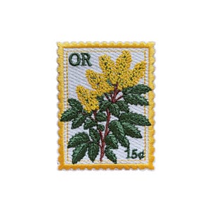 Könnte beinhalten: Gestickter Aufnäher in Form einer Briefmarke mit einer botanischen Illustration. Das Design umfasst gelbe Blüten, grüne Blätter und den Text "OR" und "15¢". Der Aufnäher hat einen weißen Hintergrund mit einem gelben Rand.