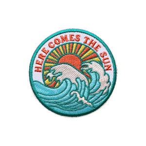 Może przedstawiać: Okrągła, haftowana naszywka z turkusową obwódką, przedstawiająca wschodzące słońce nad falami oceanu. Tekst "HERE COMES THE SUN" jest pomarańczowy. Słońce jest pomarańczowe i żółte, a fale turkusowe i białe.