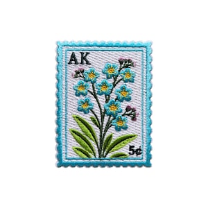 Puede incluir: Parche bordado con forma de sello postal. El parche presenta una ramita de flores azules y amarillas con hojas verdes. El texto "AK" está en la parte superior y "5¢" en la inferior. El borde es azul claro.