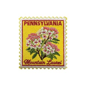 Könnte beinhalten: Gestickter Aufnäher in Form einer Briefmarke. Der Aufnäher hat einen gelben Hintergrund mit dem Text "PENNSYLVANIA" oben und "Mountain Laurel" unten. Die Mitte zeigt ein Blumendesign mit rosa und weißen Blüten.