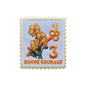 Op de afbeelding: Geborduurde patch in de vorm van een postzegel. Het ontwerp toont oranje bloemen, een sleutel en het nummer 3. De tekst "BONNE COURAGE" staat onderaan. De patch heeft een lichtblauwe achtergrond en een gele rand.