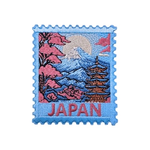Puede incluir: Parche bordado con forma de sello postal con un paisaje japonés. El diseño incluye el monte Fuji, una pagoda y flores de cerezo, con la palabra "JAPAN" en letras rosas sobre un fondo azul.