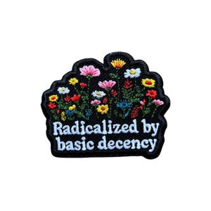 May include: Embroidered patch with a black background featuring a colorful floral design and the text "Radicalized by basic decency" in white. The flowers include pink, yellow, red, and blue blooms.