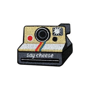 May include: Embroidered patch depicting a retro instant camera. The patch is tan, black, and white, with a rainbow stripe and the words "say cheese" embroidered in white. A fun accessory for clothing or bags.