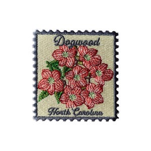 Może przedstawiać: Haftowana naszywka przedstawiająca skupisko czerwonych kwiatów derenia z zielonymi liśćmi. Naszywka ma wyhaftowane słowa "Dogwood" i "North Carolina" na kremowym tle z ciemnoniebieską obwódką.