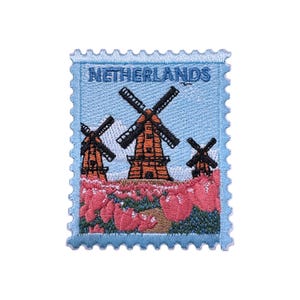 May include: Embroidered patch with a light blue background and the word "NETHERLANDS". It shows three windmills with orange bodies and black blades, set against a field of pink flowers, resembling a postage stamp.