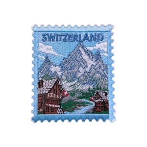 Può includere: Toppa ricamata a forma di francobollo con una scena di montagna svizzera. La toppa ha un bordo azzurro e la scritta "SWITZERLAND" in alto. L'immagine raffigura montagne innevate, due chalet e una bandiera svizzera.
